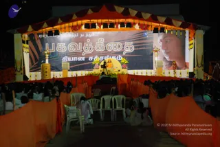 Night photo of an outdoor satsang stage under an orange and maroon canopy with yellow tassels and lights. A large banner reads Bhagavad Gita in Tamil script over a dark background with feather motifs, and a large screen on the right shows a close-up of a man with a red tilak on his forehead. On the stage, a seated figure in orange robes sits on a yellow throne with a microphone stand before him, surrounded by yellow and pink flower arrangements. Rows of white plastic chairs stand on a path lined with orange draped cloth, and a crowd of seated devotees in white fills the foreground and sides. a person: the seated figure in orange on the throne has a shaved head, but the appearance cues are limited, and the portrait on the screen is a depiction.