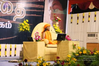 A seated figure in an orange saffron robe and orange head wrap sits on a large yellow upholstered throne-style chair on a stage. He wears a rudraksha mala around his neck and has a yellow cushion at his side. A laptop and microphone sit on a yellow draped table in front. Yellow and red flower arrangements and yellow marigold garlands decorate the stage. Behind him hangs a black banner with large tan Tamil letters and a large portrait of a man's face in profile. A white curtain and red drapery frame the backdrop, and a wall air conditioner is visible at right. One person is visible.