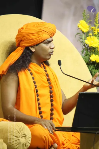 A photograph of a man seated on a large yellow cushioned chair, speaking into a small black microphone on a boom stand. He wears an orange turban with a small red mark on his forehead, an orange shawl over orange robes, and a long rudraksha mala with multiple brown beads. His long dark hair falls over his shoulders. His right hand is raised in gesture while his left rests on his lap. To the right, a black laptop sits on a table beside a vase of yellow flowers. The scene resembles a satsang seated discourse. Swamiji (probable) appears in person, identified by the orange turban, rudraksha mala and orange robes while speaking to the gathering.