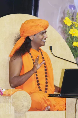 A seated man in an orange turban and orange robes, with a red bindi and a long rudraksha mala of dark beads across his chest, gestures with one raised hand while speaking. He sits on a large pale yellow throne-like chair with a microphone on a stand beside him and a laptop on a table to his right. Yellow chrysanthemums in a vase stand at the right edge against a pale backdrop. Appearance-based label: Swamiji (probable), based on the orange turban, long hair, saffron robes and rudraksha mala, and the page's note that he attended this event.