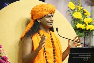 A man in saffron-orange robes and a matching orange turban speaks at a microphone during a satsang. He wears long rudraksha mala beads over his shoulders and has a red bindi on his forehead. He gestures with both hands, palms open, while smiling. Behind him is a large pale yellow circular backdrop, and yellow daisy-like flowers and pink blooms decorate the stage. A dark laptop sits at the lower right. The swami identity is not established by this image.