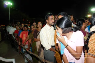 Night photograph of devotees queued along a metal railing outdoors under bright floodlights, with orange flags visible in the background. In the foreground a young woman in a pink top and dark hair tie holds a blue towel to her face, and a man in a pale yellow checked shirt and grey trousers stands at the rail beside older women in saris. Several other people line the barrier, and a white plastic chair sits at the lower left. No a person candidate is visible in the frame.