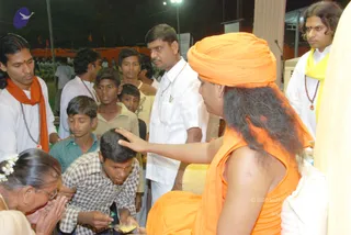 Evening outdoor darshan with a seated figure in orange clothing and a tall orange turban, hand raised in blessing over the head of a young boy in a checked shirt who bows with hands together. Around them stand and kneel devotees: a young man in white with an orange shawl and rudraksha mala, a man in white shirt with a mala, boys in casual clothes, and an elderly woman with white flowers in her hair folding her hands. A man in white with a yellow shawl and red-bead mala stands at right beside a white pillar. Orange flags and a dark night sky with stage lights appear in the background.