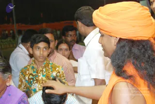 Outdoor evening darshan at night, with a dark background and an orange fence behind the crowd. In the right foreground, a man in an orange turban and sleeveless orange robe, with long, wavy dark hair, stands in profile and places his hand on the head of a seated person at lower centre, who has short dark hair. A woman in a dark-red bindi and white clothing stands nearby. Around them, devotees include a boy in a yellow, blue and brown patterned shirt gazing toward the scene, a man in a pink-striped shirt, a man in a white shirt with a gold chain, and a older woman in a purple sari at lower left. A Swamiji (probable) identification is based on the orange turban and robe with long hair, giving blessing to a devotee.