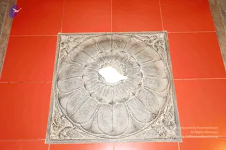 A photograph of a square grey stone slab set into an orange-red tiled floor. The slab carries a carved concentric lotus medallion with layered petals radiating from a central boss, with foliage motifs at the four corners. No people are visible; a white blank patch sits over the centre of the carving.