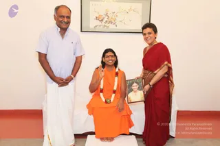 Indoor photo in a white-walled room with a framed floral painting on the wall. A seated figure in saffron-orange robes and a marigold-and-red garland sits on a white-draped platform, smiling and raising one hand in a greeting. The figure has a red bindi and long dark hair. Beside him, a man in a pale blue short-sleeved shirt and white dhoti stands with hands clasped. On the right, a woman in a deep red sari with gold border holds a framed photograph of a young woman. The seated figure is shown in person; the printed copyright text on the image is not included in the description. Three people are visible.