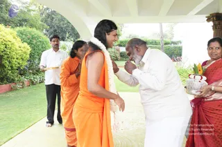 A photograph taken under a white arched veranda beside a green lawn and shrubs. A central figure with long swept-back dark hair wears a saffron-orange sleeveless robe and a long white flower garland over the shoulders, smiling as an older man in white kurta and dhoti bends forward to offer respect with folded hands. A second figure in orange robes stands bowed behind them. On the right, a woman in a red sari holds a silver tray with flowers and a white basket-shaped offering. A man in a white shirt and black trousers stands at left holding a gold-edged plate.