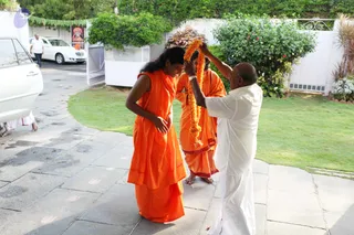 Outdoors on a stone-paved path beside a green lawn, a man in white dhoti and shirt places an orange marigold garland over the head of a figure in saffron robes with long dark hair. A second person in orange robes stands behind them, barefoot. Parked white cars, a flowering hibiscus bush and a white boundary wall are in view, and a framed portrait of a saffron-clad figure with a crown-like head covering hangs on a stand in the background. The garlanding appears to be a welcome ritual. The saffron-robed figure with long swept-back hair and orange robes matches the page's Swamiji cues, so this is a probable in-person appearance; the page places him at this house visit.