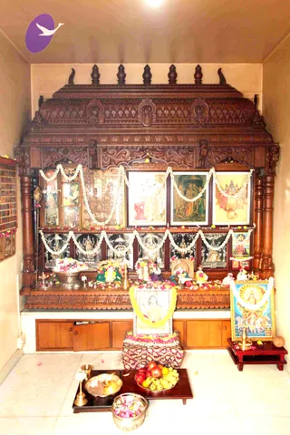 Photograph of a home shrine room with an ornately carved dark wooden altar cabinet against a pale wall. Framed printed deity pictures hang above a row of small silver murtis, and the shelves are decorated with white jasmine garlands and fresh flowers. Brass lamps, a brass kalasha-style vessel, and a bowl of petals sit on the altar. On the floor in front a tray holds a brass lamp, a brass bell, and a plate of apples, bananas and mangoes, with a flower-filled bowl beside it. A framed picture of a sun-bearing deity rides a chariot pulled by horses at right. No people are visible.