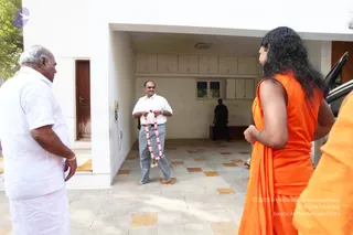 A photograph of a house visit at a white building entrance with tiled floor. In the centre, a man in a white shirt and grey trousers holds a pink flower garland. At left, an older man in white shirt and white dhoti faces him. At right, a long-haired figure in a saffron-orange sleeveless robe with a draped orange shawl stands with their back to the camera, next to another person in orange at the far right edge. A figure in dark clothing stands in the background doorway, and a car is partly visible at right. a person cue is the long swept-back hair and saffron robe on the back-turned figure, but the face is not visible, so the identity remains uncertain.