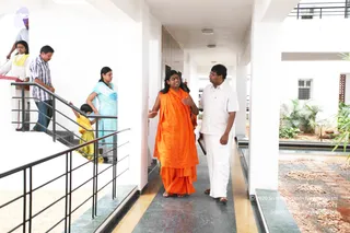 A photograph in a white, open-air corridor with a grey-green marble floor and a courtyard garden of palms and pebbles on the right. In the centre, a man with long dark swept-back hair wears a saffron-orange robe and gestures with one hand while talking. He appears to be a person, with long swept-back hair and saffron clothing, and is walking beside a man in a white shirt and white dhoti who holds a folder. Behind them on a staircase at left, several people in white, yellow and blue clothes walk up and down, including a woman in a light blue sari and a child in yellow.