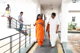 A photograph in a white-walled corridor of a modern building with black metal railings and a grey-and-yellow tiled floor. A figure with long dark hair in a saffron-orange shawl and orange garment walks barefoot, gesturing with one hand while speaking. Beside them a man in a white short-sleeved shirt and white dhoti walks barefoot. Behind them another person in orange carries a folder. On the left, a person in white with a yellow shawl stands near the staircase, with two other people in the background, one in a striped shirt on the stairs. Through the right-side opening a courtyard shows potted palms and gravel paving.