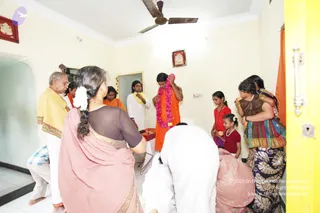 Indoor photo of a house visit in a small room with cream walls, a white ceiling fan and a framed deity picture on the wall. A bare-chested figure in an orange-saffron cloth stands centre, wearing a long pink-and-white flower garland and holding a garland in his hands; a person candidate by saffron wrap and long swept hair, though uncertain. Around him stand a man in a white dhoti with a yellow-orange shawl at left, a man in white with a yellow shawl beside him, a woman in a pink saree with a white flower in her hair in the foreground, two girls in red tops, and a woman in a dark patterned saree holding a child. A person in white bends forward at the bottom centre near a red cloth with offerings on a low table.