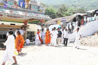 A daytime outdoor scene at a South Indian temple entrance with a colourful gopuram-style facade painted with figures and turquoise, pink and yellow ornamentation. A group walks down a dusty dirt path past a white staircase, and many people in white dhotis and shirts line the steps and walk along the road. In the centre, a figure in an orange saffron robe with a green-and-orange garland, bare-footed, is flanked by people in white. Another person in orange robes walks at left, and a figure in white with a red-orange shawl walks near the centre. Several people carry steel vessels and buckets near the temple wall, and a hillside rises in the background.