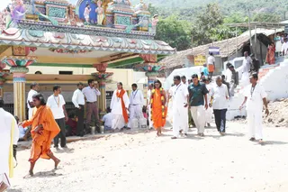 Outdoor street scene in front of a colourfully painted temple gopuram-style entrance with columns and sculpted figures above. A woman in a saffron-orange robe with a garland of mixed flowers walks barefoot on a dusty dirt road, flanked by men in white dhoti-and-shirt attire, several with lanyards. Another barefoot figure in orange robes walks at the left edge. Behind them, a man in a white shirt and a man in a dark green shirt walk, a few onlookers stand near steps, and a tiled-roof building and hillside trees appear in the background. Several plastic containers sit on the ground near the temple steps.
