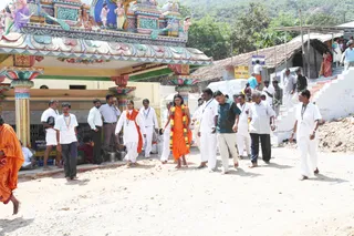Outdoors on a dusty dirt road beside a colourful gopuram-style temple entrance with painted deity figures and pillars. A central figure in a saffron-orange robe, with long dark hair and a marigold garland over the shoulders, walks barefoot in the middle of a group. Around them men in white shirts and dhotis, some with lanyards, and a man in a green shirt walk alongside, with a woman in white and an orange shawl nearby. Bystanders stand under the temple roof and on steps at right, and a tiled-roof building and hillside trees sit in the background.