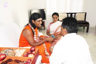 A seated man in an orange sleeveless sannyasi garment with a long flowing wrap and a garland of white flower strands is smiling and handing a small card or envelope to a man in a white shirt who leans forward with hands together. Shoulder-length dark hair and a white tilak on the forehead are visible on the seated figure, who wears a garland of onion-like pale bulbs. Beside them a woman in a white and pink sari with a bindi sits smiling on a wooden bench near a television and white curtains. In the foreground a wooden tray holds red apples, wrapped sweets, and strings of rudraksha-like beads. Presentation of the seated figure's cues is consistent with a person candidate.