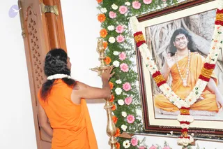 A person in an orange sannyasi-style garment with long dark hair and a white garland stands at a wall, holding a brass oil lamp (aarti lamp) toward a framed portrait. The framed portrait, labelled with a printed copyright line, shows a smiling man in orange robes with a white bead necklace seated before a banyan tree trunk, one hand raised in blessing. The portrait is decorated with a garland of white, pink and red flowers, orange gerbera blooms and fern leaves. The scene suggests a devotional lamp offering before a photograph. The orange-robed figure is a person candidate by appearance: long swept hair and saffron clothing, with a white flower garland, though the face is turned away.