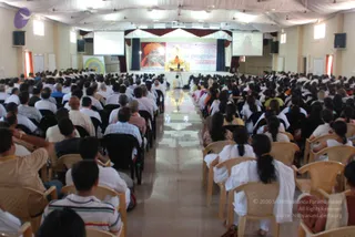A large indoor hall filled with seated attendees in rows of plastic chairs, facing a raised stage at the far end. Many attendees wear white clothing, and some are in light shirts and kurtas. The stage backdrop shows a large banner reading part of a program title, with a framed portrait of a saffron-robed figure with long hair and a crown-like headdress on the left panel and a central image of a seated figure before a lamp arrangement. Projection screens hang above the stage, and a shrouded white canopy overhead drapes the ceiling. A small figure stands near the stage front. The floor is polished and reflective. The scene shows a seminar-style session with an audience facing the stage.