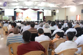 A large audience of seated attendees, mostly men in white shirts and some in striped or dark clothing, faces a stage at a hall for an NSP program. On the stage a figure in orange and gold sits on a golden throne, with a large portrait on the left backdrop showing a man in an orange turban. Projection screens flank the stage, and white draped fabric covers the ceiling. Plastic chairs fill the room, and a young child in orange sits among the audience at right.