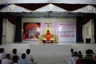 A photograph of a stage event in a hall with a white and maroon draped ceiling valance and dark side curtains. A seated figure in an orange-saffron robe with hands pressed together in prayer sits on an ornate gold throne with a gold canopy and carved gold side panels, centered on a white raised platform. Behind the throne hangs a large banner showing a smiling man in an orange turban with a red tilak, labelled as a portrait captioned Paramahamsa Nithyanan[anda], next to a banner reading Nithyananda Dhyanapeetam and a pink-and-grey banner reading program with Level 2. Two standing fans flank the platform. Around ten to twenty seated audience members in casual clothes, mostly viewed from behind, face the stage, with a few seated at the front left and right edges. a person is seated on the throne in saffron, with a visible tilak and his hands in prayer, and an ornate gold crown-like canopy above.