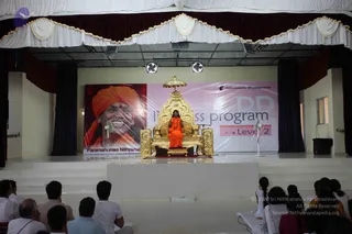 A seated figure in orange robes sits on an ornate gold throne with a canopy on a raised white stage in an indoor hall. The seated figure holds hands together in front of the chest and wears a rudraksha mala. Behind the throne hangs a large banner showing a smiling man in an orange turban with the printed caption "Paramahamsa Nithyanand..." on the left, alongside the text "Nithyananda Dhyanapeetam" and "Level 2" on a pink panel. Floor fans stand on either side of the stage. Several people in white and grey clothes sit facing the stage in the foreground, and a few women in light saris sit at the right edge.
