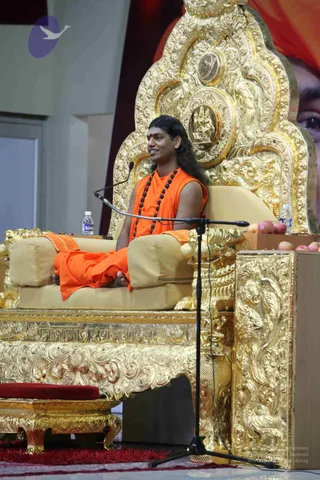 A photograph of a man seated cross-legged on an ornate gold throne with carved floral and figural relief, beside a microphone stand. He wears an orange saffron robe draped over one shoulder and a long rudraksha mala of dark beads, with a red bindi on his forehead, and sits with a small orange cloth on the armrest. Water bottles and a few apples rest on a gold-carved side table, and a gold footstool with a red cushion sits in the foreground. A large backdrop behind shows a partial portrait with a red turban. The figure appears to be Swamiji (probable), given the saffron robe, long swept-back hair, rudraksha mala and the throne setting, and the event page flags his presence. A single person is visible.
