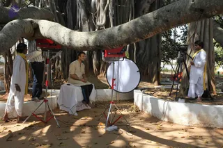 A film crew records an interview beneath a large spreading banyan tree with aerial roots and a long horizontal limb. A man in a beige kurta and dark trousers sits barefoot on a white-draped platform, facing the camera. Two red lighting units on stands flank him, with a white reflector disc in front of the right light. On the left, a crew member in white kurta and dhoti with a yellow shawl stands on the low white wall, and on the right another white-clad person with a yellow shawl operates a camera on a tripod. Dry leaves cover the ground and the wall edges are whitewashed.