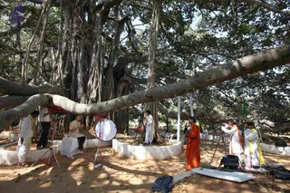 A photograph of a film crew shooting an interview under a large banyan tree with many aerial roots and a long horizontal limb. A seated man in a cream kurta and light trousers sits on a white-draped chair in the foreground, facing a second man with a handheld object. Around him, a man in a white kurta with a yellow shawl stands near a camera tripod, and a man in a saffron-orange robe stands on the right beside a woman in a white outfit with an orange shawl adjusting a camera. Other crew members in white and yellow shawls work nearby. Two red lighting units on stands and a round white reflector are set up, with equipment cases on the sandy ground and white-painted stone platforms around the tree.