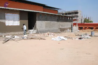 A dusty open construction site in daylight beside a partly built concrete block with grey plastered walls, an orange-painted brick wall, and a corrugated green roof edge. A man in a white shirt and grey trousers stands at the building entrance near a printed sheet taped to the wall. Stacked wooden poles, loose bricks, white cloth, sacks, a metal drum and buckets lie on the sand. Behind, a multi-storey concrete frame with scaffolding and tall construction rods rises under a clear sky. No ritual or a person presence is visible.