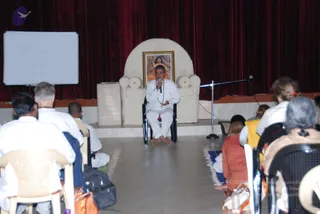 A seated man in an all-white outfit speaks into a handheld microphone on a raised stage in a hall with red velvet drapes. He sits on a black chair before a cream arched throne-like seat. Above the seat hangs a framed portrait of a figure in saffron and orange robes. Barefoot, he holds a rudraksha-style beaded mala around his neck. A whiteboard with hand-drawn circles stands at the left. In the foreground, audience members sit on white plastic chairs, some in white clothes and one woman in orange, with bags on the floor.