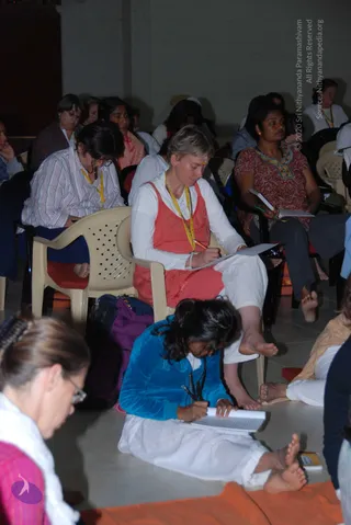 Photo of a seated audience at a person Spurana Convention session in a plain hall with whitewashed walls. Foreground: a woman in a blue velvet top and white trousers sits cross-legged on an orange cloth, writing in a notebook. A woman in a coral sleeveless top over white clothes sits on a white plastic chair, writing on a pad and wearing a yellow lanyard. Behind them, women in striped shirts, a red patterned kurta, and other seated attendees take notes. No a person figure is visible; no speaker is shown on stage.