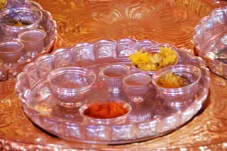 A close view of ceremonial offering vessels on a brass tray. A large scalloped clear glass plate holds several small glass bowls: one with orange-red kumkum or powder, one with yellow marigold-type flowers, and others empty. Behind it are more brass trays with engraved floral patterns and additional glass bowls containing yellow flowers. No people are visible; the arrangement suggests items prepared for a puja offering.