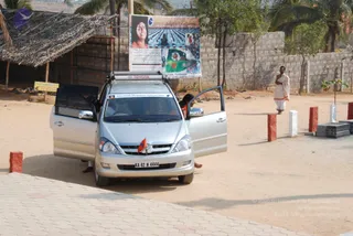 A silver Toyota minivan with its front and rear side doors open is parked on a sandy compound beside a paved walkway. A small saffron flag is mounted on the front grille and the windshield shows a sticker reading Nithyananda Dhyanapeetha Mahasamsthana. A person in orange is partly visible stepping out of the rear door. Behind the van, a roadside board shows a portrait labelled Nithyananda Dhyanapeetha and a photo of a crowd. A man in white clothes with an orange shawl walks along the right side near small red and white pillars. A thatched shelter and a block wall with palm trees stand in the background.