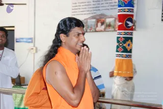 A photograph of a man with long, swept-back black hair and a small red bindi, wearing a sleeveless saffron-orange robe, standing with palms pressed together in namaste and smiling. Behind him stands a second person in an orange robe, partly visible, and to the right a person in white dhoti and shirt. A tall pillar wrapped in a multicoloured cloth with flower and bird motifs and an orange base stands beside him. Framed pictures hang on the white wall behind, and a steel railing runs across the lower frame. a person: the central man has long swept-back hair, saffron clothing and a bindi, consistent with the page's claim of his presence at this event.