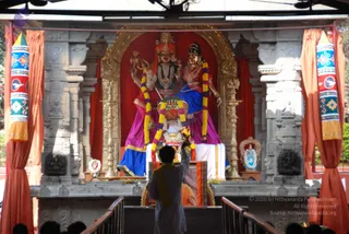 A photograph of a temple shrine with a large deity murti of a standing figure in red and gold, flanked by a second figure in blue and magenta draped garments, both adorned with garlands of yellow and orange flowers. In front, a figure in a grey kurta with an orange shawl stands with raised arm, facing the shrine. Dark-haired devotees are visible at the bottom edge of the frame. Orange curtains, ornate stone pillars, decorative banners with blue and red patterns, and brass oil lamps frame the altar.