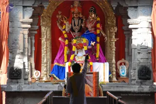 A photograph of a temple shrine with a large deity murti pair in the centre: a male figure with a tall red and gold crown, holding a trident-like staff, and a female figure in a blue and magenta sari with gold jewellery, both draped in marigold and yellow garlands. A ceremonial shrine sits in front with a decorated kalasha-like object and a flower garland. Ornate grey stone pillars frame the scene with red curtains and brass oil lamps on either side. In the foreground, one person in a white shirt and saffron-yellow shawl stands at the rail with an arm raised toward the murti, appearing to perform worship or a blessing gesture. A faint portrait of a robed figure in orange appears on the lower altar area.