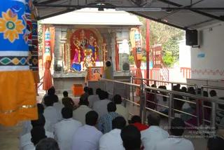 A temple shrine under a corrugated roof with orange and multicoloured draped banners. A priest in a white garment and saffron-yellow shawl stands at a central altar before a deity murti decorated with garlands, with a framed portrait placed on the altar. Thirty or more devotees, mostly men in white and light shirts with some women in saffron and pink saris, sit in rows on the floor facing the shrine behind metal railings. A brick-red and white temple structure with a small blue sign is visible at right.