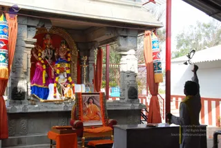 A photograph of a temple shrine with a stone shrine structure framed by orange curtains and striped banners. Inside, deity murti of a goddess figure with red and gold ornaments and a standing deity with a sword, garlanded with marigold and white flower garlands, sit on a white-draped platform. In front, a framed poster portrait of a man in orange robes and a rudraksha mala sits on a red and gold chair draped with an orange cloth. A man in white clothes with an orange shawl stands at the right, raising a bell or lamp toward the shrine. Brass lamps and a small offering tray are visible.