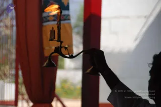 Close-up photograph of a brass aarti lamp with a lit flame held in a silhouetted hand at the right edge. The lamp has a curved handle and multiple tiers, with a burning flame rising from the top. Behind it hang red curtains and a yellow-and-patterned cloth, with a white wall and red pillar visible. The figure is in shadow, showing only a sleeved white arm and a wrist band, so no face is visible. The background shows a doorway opening onto blurred greenery.