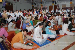 A large indoor hall filled with seated attendees, mostly women and some men, on orange and blue mats. Most wear white or pastel clothing, with orange, green, blue and pink garments scattered among them; several wear rudraksha-style malas and white scarves. Some hold notebooks or brochures. Water bottles and bags sit on the floor, and a white-clad woman in the foreground looks forward while others sit attentively. The hall has white stage rigging, colourful banners on the pillars and wooden panels, and ceiling fans. A blue mat reads partly as NITHYANANDAM in Roman letters, and the lower right shows a red-brown sitting mat with a tan bag. No central figure is clearly identifiable in the visible frame.