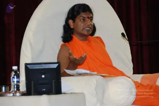 A man seated on a large white cushioned chair speaks while gesturing with his right hand open, palm up. He wears an orange saffron sleeveless garment, has long swept-back dark hair and a red bindi on his forehead. A black microphone stands at right against maroon velvet drapes. In front of him are a black monitor, a clear water bottle and a small silver bowl with a few items, and papers rest on his lap. The setting appears to be a seated discourse or session.