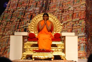 A seated man in an orange-saffron robe sits on an ornate gold throne with a large circular golden backrest and red cushions, hands pressed together in anjali mudra at chest level with eyes closed. Behind him is a large backdrop printed with colourful temple gopurams and sculpted figures. A white side table with a small object sits to his left, and the throne rests on a golden plinth with a red cushion. The figure has long swept-back dark hair and a bare chest visible above the orange cloth, a combination of cues consistent with the page's a person label, so this is a probable in-person appearance. The framing is a wide stage view with no other people visible.