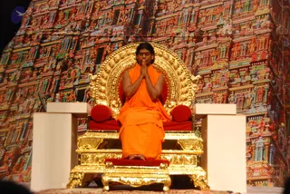 A seated man in saffron-orange robes sits on an ornate gold throne with red velvet cushions, palms pressed together in namaste with eyes closed. A decorative backdrop of a colourful multi-tiered temple gopuram with painted deity figures fills the background. The throne is flanked by two white pedestals, and a red-cushioned footstool supports the feet.