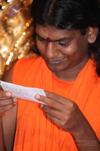 Close-up photo of a smiling man in an orange-saffron robe with long dark hair and a red tilak on his forehead, looking down and reading a small handwritten note held in both hands. Behind him is a blurred golden, sparkling backdrop. The handwritten note is partially legible. A purple logo appears at upper left, and copyright text overlays the lower right corner.