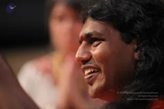 A close-up photograph of a smiling man with long, dark, swept-back hair and a red tilak mark on his forehead, wearing orange-red clothing. He is turned to his left, mouth open in a broad smile. Behind him and out of focus, a woman with her hands pressed together in a namaste gesture wears a white garment. The background is dark and blurred. The image is watermarked at the lower right and corner logo, which are not described.