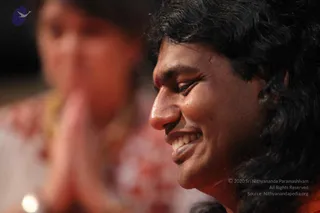 Close profile photograph of a smiling man with long dark wavy hair and a small red kumkum mark on his forehead, eyes lowered, shown in warm reddish light. Behind him and out of focus, a woman in a white and red patterned garment holds her palms pressed together in anjali mudra. The man's dark hair and smile are the clearest features, and the background is a blurred gathering of devotees at a darshan-style event.