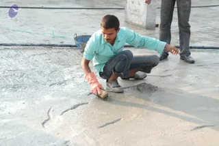 A construction worker in a light turquoise button-up shirt and dark jeans crouches on a freshly poured concrete slab, brushing its surface with a wooden-handled brush while wearing orange rubber gloves. A blue metal bucket and green cable lie nearby, and another worker's legs in grey trousers and dark shoes stand at the top right beside a concrete column. No a person candidate is visible; the scene shows site work.