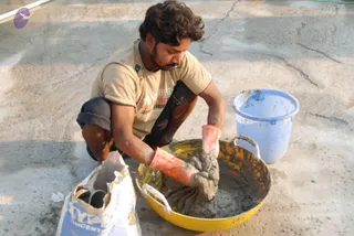 A worker crouches on a concrete surface, mixing wet cement in a yellow plastic basin with orange rubber gloves. He wears a beige t-shirt with printed lettering, dark trousers and has short curly hair and stubble. Beside him are an open cement bag with a blue and white label and a light blue bucket. The scene looks like construction work at the site.