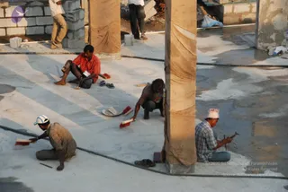 A construction site photo of workers finishing a concrete floor between tan cloth-wrapped pillars. Four men work at ground level: one in a red shirt sits on his heels, one in a grey shirt crouches with a red-bristled brush, one in a checked shirt and white cap sits with a hammer and chisel, and one in a checked shirt and white cap brushes wet concrete. Lengths of black hose run across the floor, and cinder blocks and stacked materials are visible behind them. The scene is a building under construction.